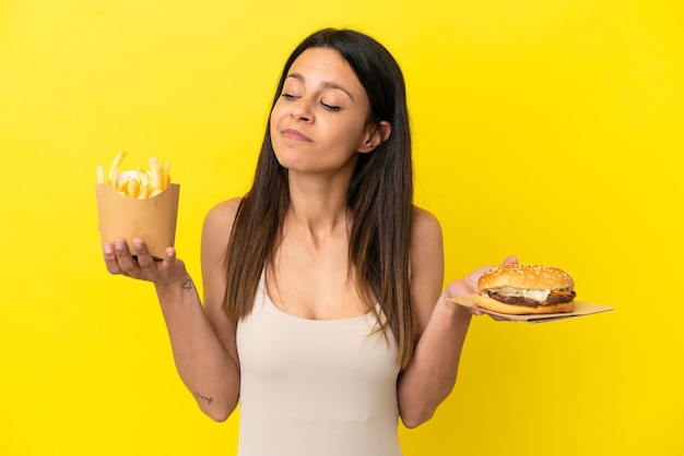 Jovem mulher caucasiana segurando hambúrguer e batatas fritas isoladas em um fundo amarelo