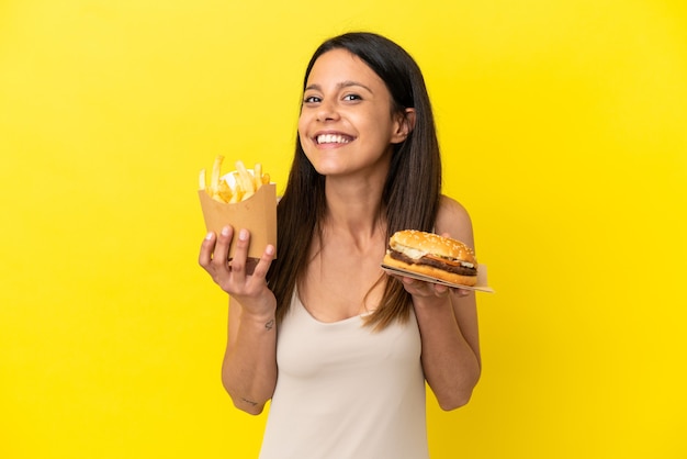 Jovem mulher caucasiana segurando hambúrguer e batatas fritas isoladas em um fundo amarelo