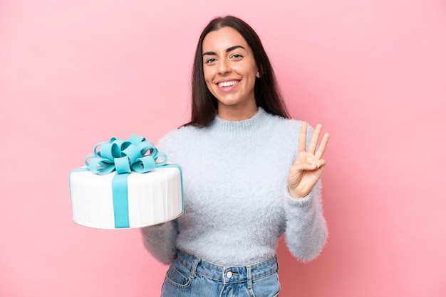 Jovem mulher caucasiana segurando o bolo de aniversário isolado no fundo rosa feliz e contando três com os dedos