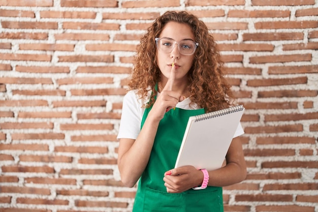 Jovem mulher caucasiana segurando o caderno de arte pedindo para ficar quieto com o dedo nos lábios silêncio e conceito secreto