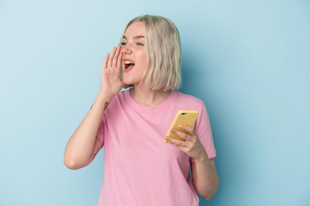 Jovem mulher caucasiana, segurando o celular isolado no fundo azul, gritando e segurando a palma da mão perto da boca aberta.