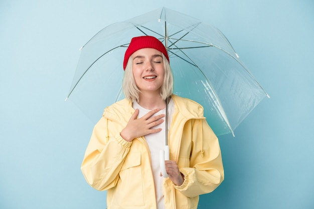 Jovem mulher caucasiana, segurando o guarda-chuva isolado no fundo azul, ri alto, mantendo a mão no peito.