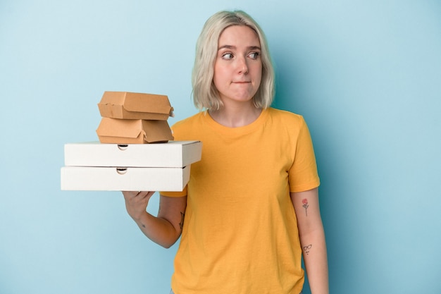 Jovem mulher caucasiana segurando pizzas e hambúrgueres isolados em um fundo azul confusa, sente-se duvidosa e insegura.