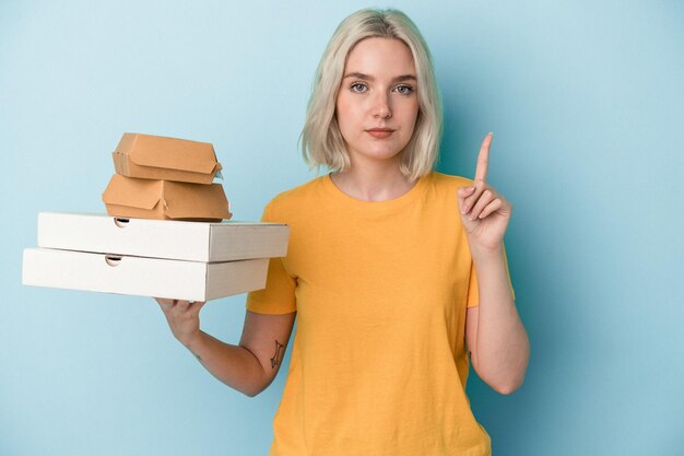 Jovem mulher caucasiana segurando pizzas e hambúrgueres isolados sobre fundo azul, mostrando o número um com o dedo.