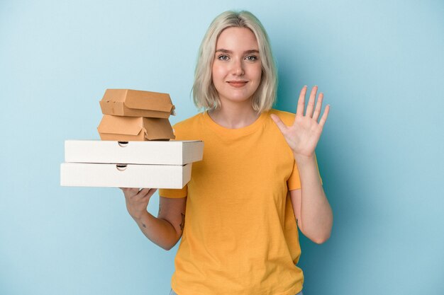 Jovem mulher caucasiana, segurando pizzas e hambúrgueres isolados sobre fundo azul, sorrindo alegre mostrando o número cinco com os dedos.