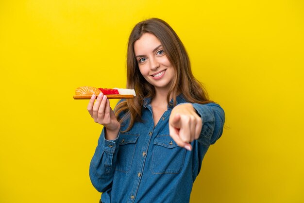 Jovem mulher caucasiana segurando sashimi isolado em fundo amarelo aponta o dedo para você com uma expressão confiante