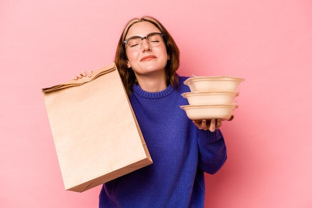 Jovem mulher caucasiana segurando tupperware e saco de viagem isolado no fundo rosa