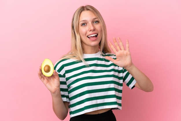 Jovem mulher caucasiana segurando um abacate isolado em um fundo rosa e saudando com a mão com uma expressão feliz