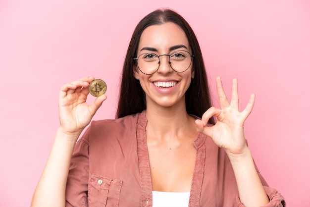 Jovem mulher caucasiana segurando um Bitcoin isolado no fundo rosa mostrando sinal de ok com os dedos