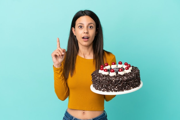 Jovem mulher caucasiana segurando um bolo de aniversário isolado em um fundo azul com a intenção de perceber a solução enquanto levanta um dedo