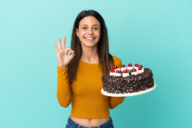 Jovem mulher caucasiana segurando um bolo de aniversário isolado em um fundo azul, mostrando um sinal de ok com os dedos