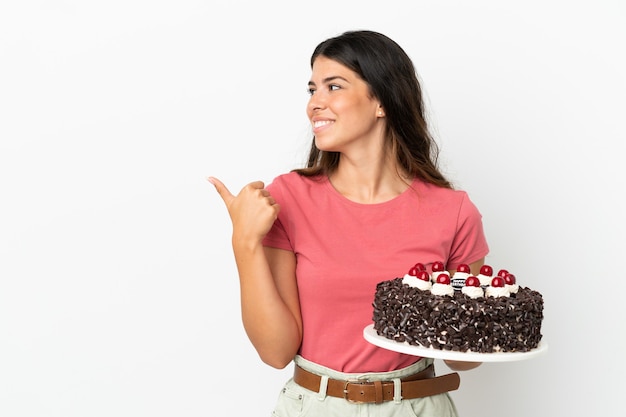 Jovem mulher caucasiana segurando um bolo de aniversário isolado no fundo branco apontando para o lado para apresentar um produto