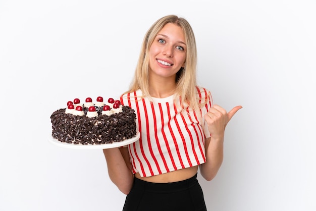 Jovem mulher caucasiana segurando um bolo de aniversário isolado no fundo branco apontando para o lado para apresentar um produto