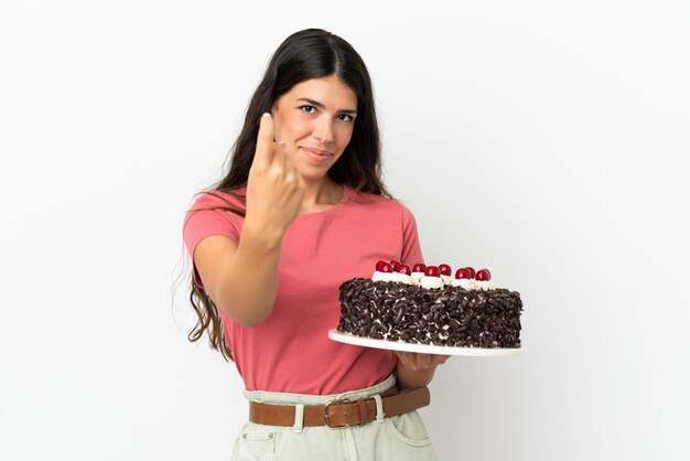 Jovem mulher caucasiana segurando um bolo de aniversário isolado no fundo branco fazendo um gesto de vir.