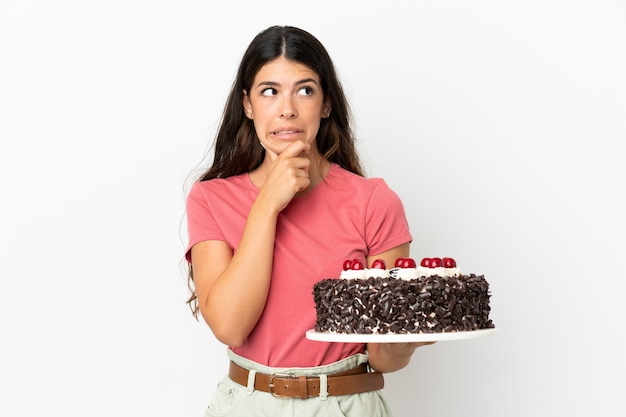 Jovem mulher caucasiana segurando um bolo de aniversário isolado no fundo branco, tendo dúvidas e pensando