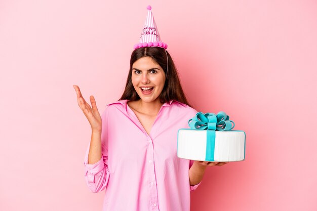 Jovem mulher caucasiana segurando um bolo para comemorar um aniversário isolado na parede rosa, recebendo uma agradável surpresa, animada e levantando as mãos.