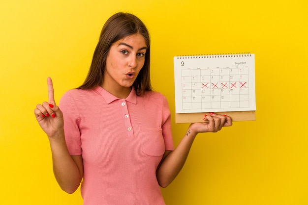 Jovem mulher caucasiana, segurando um calendário isolado em fundo amarelo, tendo uma ótima ideia, o conceito de criatividade.