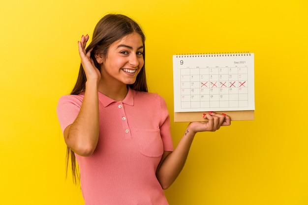 Jovem mulher caucasiana segurando um calendário isolado em fundo amarelo, tentando ouvir uma fofoca.