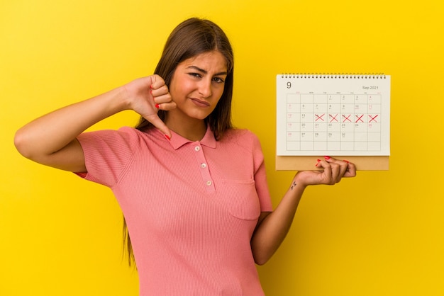 Jovem mulher caucasiana segurando um calendário isolado em um fundo amarelo, mostrando um gesto de antipatia, polegares para baixo. Conceito de desacordo.