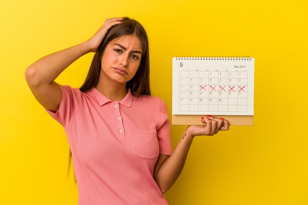 Foto jovem mulher caucasiana, segurando um calendário isolado em um fundo amarelo, sendo chocada, ela se lembrou de uma reunião importante.