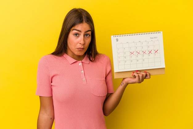 Foto jovem mulher caucasiana segurando um calendário isolado no fundo amarelo encolhe os ombros e abre os olhos confusos.