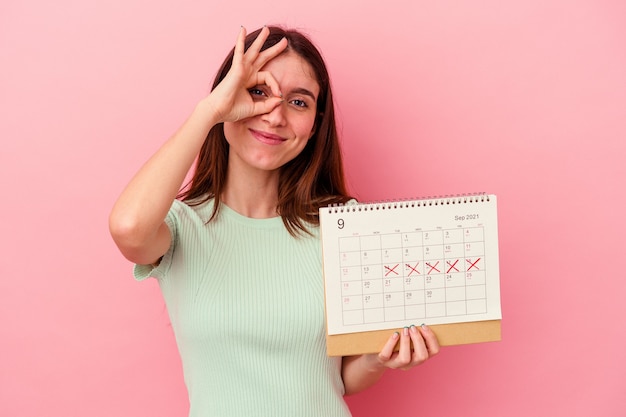 Foto jovem mulher caucasiana, segurando um calendário isolado no fundo rosa, animado, mantendo o gesto ok no olho.