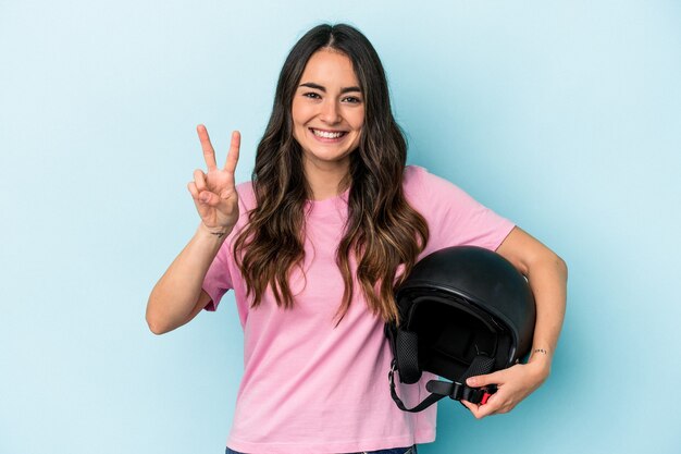 Foto jovem mulher caucasiana segurando um capacete de motocicleta isolado em um fundo azul, mostrando o número dois com os dedos.