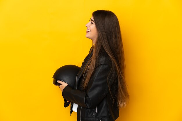 Jovem mulher caucasiana segurando um capacete de motociclista isolado em um fundo amarelo, rindo na posição lateral