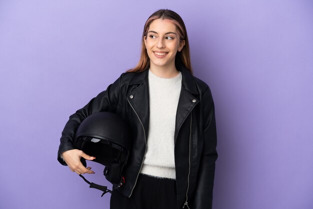 Jovem mulher caucasiana segurando um capacete de motociclista isolado em um fundo roxo, tendo uma ideia enquanto olha para cima