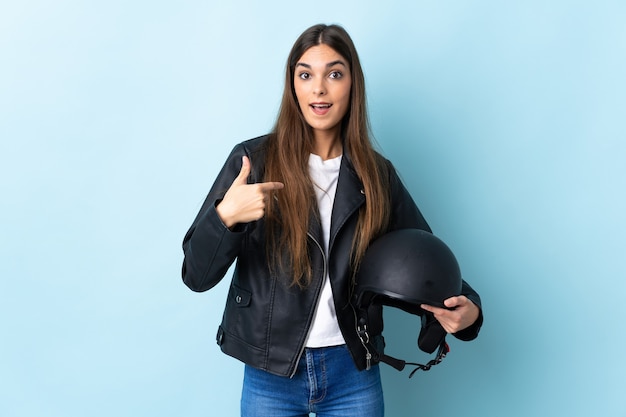 Jovem mulher caucasiana segurando um capacete de motociclista isolado em uma parede azul com expressão facial surpresa