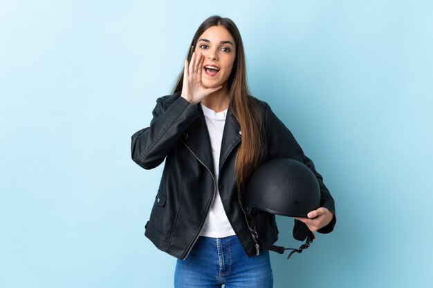 Jovem mulher caucasiana segurando um capacete de motociclista isolado na parede azul, gritando com a boca aberta