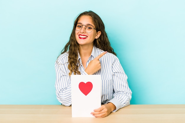 Jovem mulher caucasiana, segurando um cartão de dia dos namorados isolado, sorrindo e apontando de lado, mostrando algo no espaço em branco.