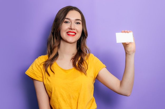 Foto jovem mulher caucasiana segurando um cartão na mão e sorrindo, vestindo uma camiseta amarela sobre um fundo lilás no estúdio. crédito online. brincar