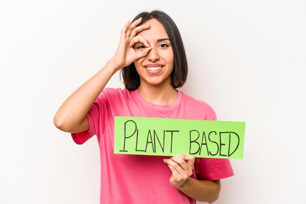 Jovem mulher caucasiana segurando um cartaz baseado em plantas isolado no fundo branco animado mantendo o gesto ok no olho