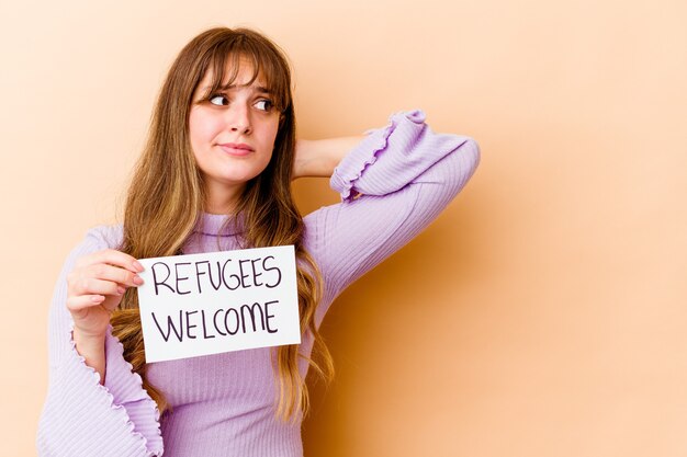 Jovem mulher caucasiana segurando um cartaz de boas-vindas aos refugiados isolado tocando a parte de trás da cabeça, pensando e fazendo uma escolha.