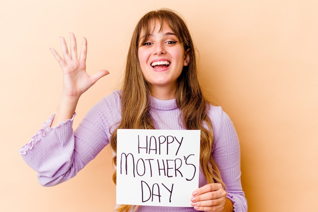 Jovem mulher caucasiana, segurando um cartaz de feliz dia das mães isolado, recebendo uma agradável surpresa, animada e levantando as mãos.