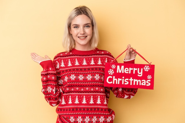 Jovem mulher caucasiana segurando um cartaz de feliz Natal isolado em fundo amarelo, mostrando um espaço de cópia na palma da mão e segurando a outra mão na cintura.