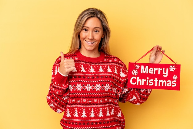 Jovem mulher caucasiana segurando um cartaz de feliz Natal isolado em um fundo amarelo, sorrindo e levantando o polegar