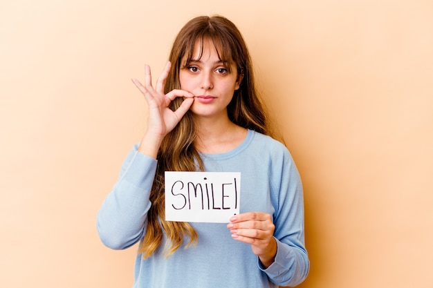 Jovem mulher caucasiana, segurando um cartaz de sorriso isolado com os dedos nos lábios, mantendo um segredo.