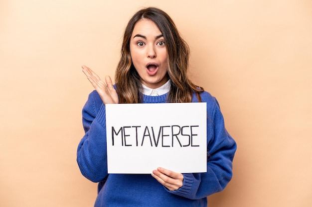 Jovem mulher caucasiana segurando um cartaz do metaverso isolado em fundo bege surpreso e chocado