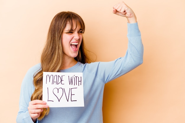 Jovem mulher caucasiana segurando um cartaz feito com amor isolado levantando o punho após uma vitória, o conceito de vencedor.