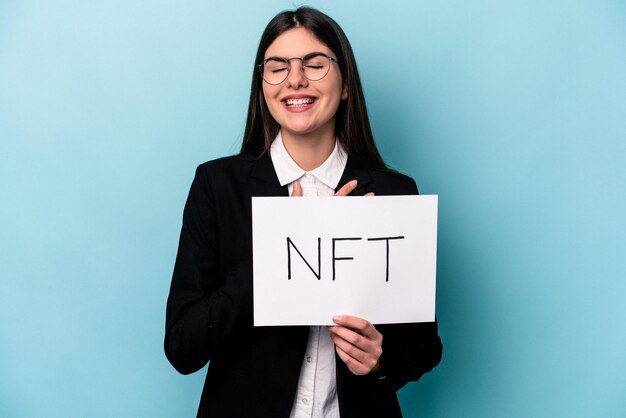 Jovem mulher caucasiana segurando um cartaz NFT isolado em fundo azul ri alto, mantendo a mão no peito