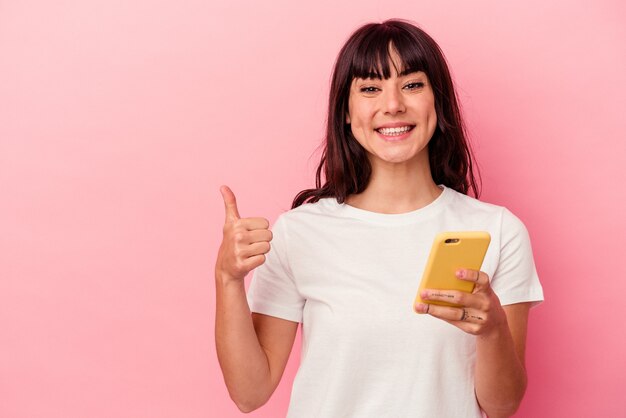 Jovem mulher caucasiana segurando um celular isolado no fundo rosa, sorrindo e levantando o polegar