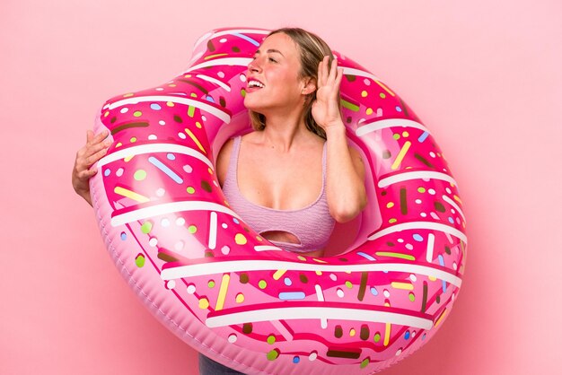 Jovem mulher caucasiana segurando um colchão de ar isolado no fundo rosa tentando ouvir uma fofoca