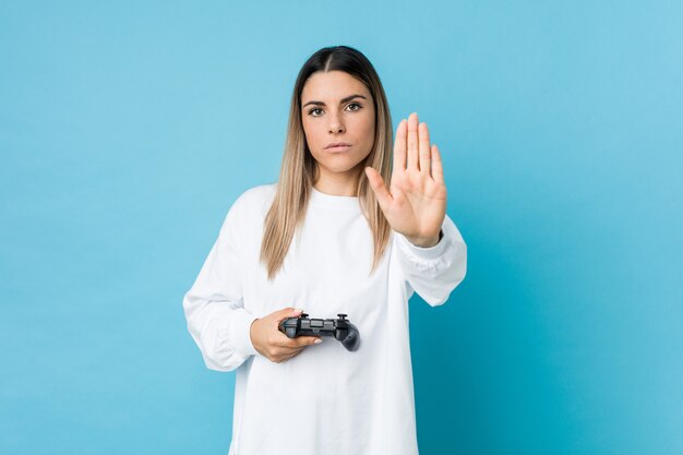 Jovem mulher caucasiana, segurando um controlador de jogo em pé com a mão estendida, mostrando o sinal de stop, impedindo-o.