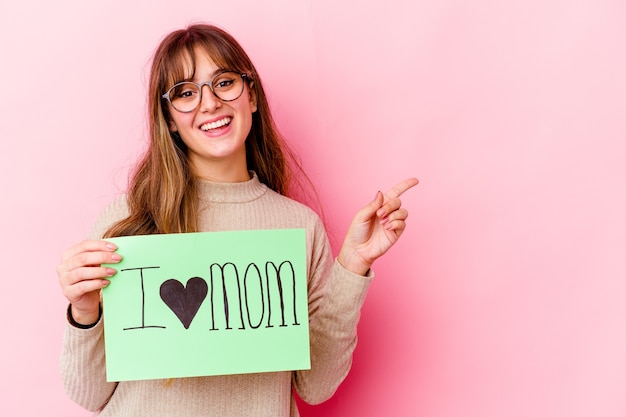 Jovem mulher caucasiana segurando um Eu amo a mãe isolada sorrindo e apontando de lado, mostrando algo no espaço em branco.