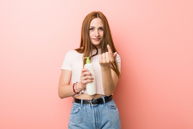 Jovem mulher caucasiana, segurando um frasco de creme, apontando com o dedo para você, como se convidando se aproximar.