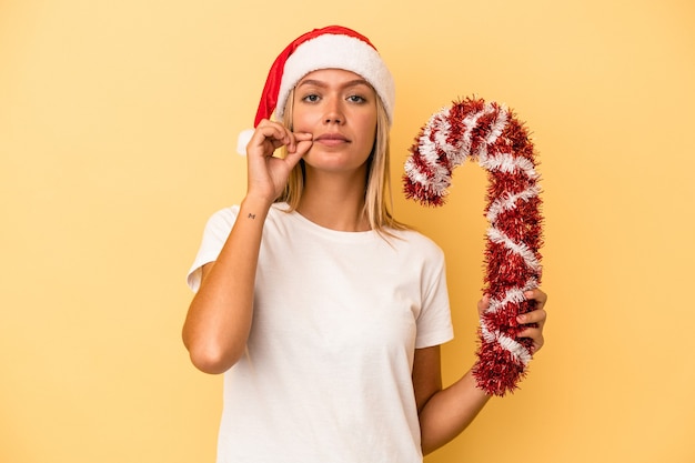 Jovem mulher caucasiana, segurando um grande bastão de Natal, isolado em um fundo amarelo, com os dedos nos lábios, mantendo um segredo.