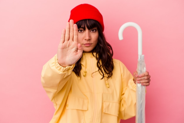 Jovem mulher caucasiana, segurando um guarda-chuva isolado em um fundo rosa em pé com a mão estendida, mostrando o sinal de stop, impedindo-o.