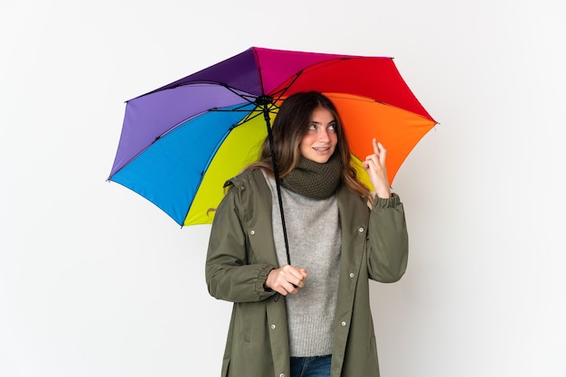 Jovem mulher caucasiana segurando um guarda-chuva isolado na parede branca, cruzando os dedos e desejando o melhor